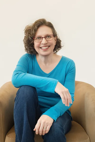 Leaf, smiling, seated in a tan armchair in a blue shirt and jeans, one arm resting across a knee. A white woman with chin-length curly brown hair and glasses.